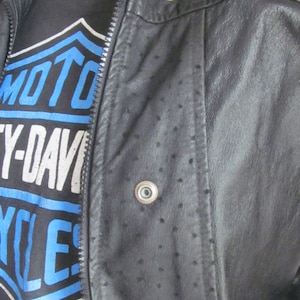 May include: A black leather jacket with a zipper closure and a dotted pattern. The jacket has a stand-up collar and a snap button closure at the neck. The jacket is worn over a black t-shirt with a blue and white graphic that says "MOTO CY-DAVI YCLES".