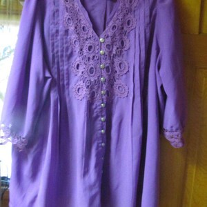May include: A long-sleeved, button-down purple blouse with a V-neck. The blouse has a detailed, open-weave lace yoke and cuffs, and vertical pleats on the front. Small, round buttons fasten the front. The sleeves are three-quarter length.