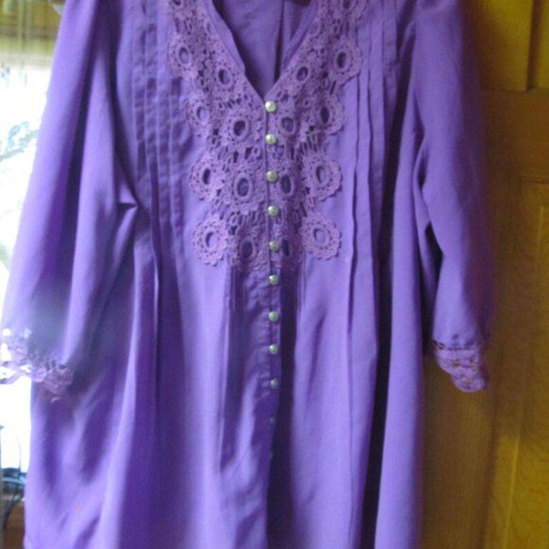 May include: A long-sleeved, button-down purple blouse with a V-neck. The blouse has a detailed, open-weave lace yoke and cuffs, and vertical pleats on the front. Small, round buttons fasten the front. The sleeves are three-quarter length.