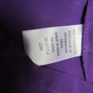 May include: A close-up shot of a purple garment with a white label. The label states "100% Polyester Exclusive of Decoration" and includes details such as "Made in India", "RN 88842", and size "22W".