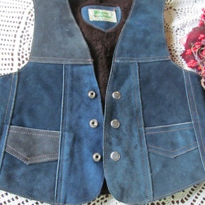 Puede incluir: Un chaleco de gamuza azul con detalles en cuero marrón y forro de piel sintética marrón. El chaleco tiene cierre de presión y varios bolsillos. La etiqueta dice "Genuine Leather".