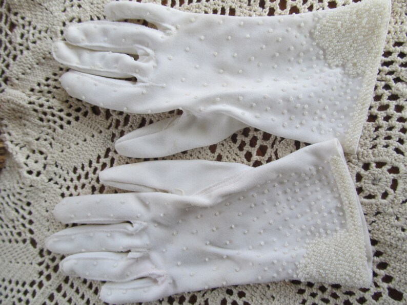 May include: A pair of white cotton gloves with a dotted pattern. The gloves have a beaded trim around the wrist.