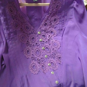 May include: A lilac blouse featuring a V-neck and a decorative crochet design around the neckline. The blouse has small, round buttons down the front and is made of a lightweight fabric.