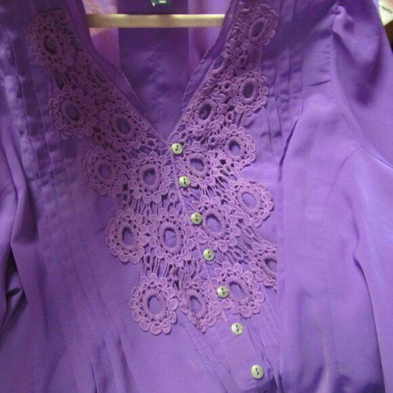 May include: A lilac blouse featuring a V-neck and a decorative crochet design around the neckline. The blouse has small, round buttons down the front and is made of a lightweight fabric.