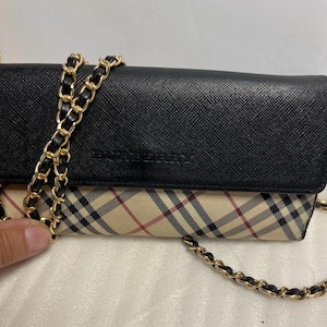 May include: A black and beige crossbody bag with a gold chain strap. The bag features a classic plaid pattern on the bottom half and a solid black top. The brand name "BURBERRY" is subtly embossed on the front.