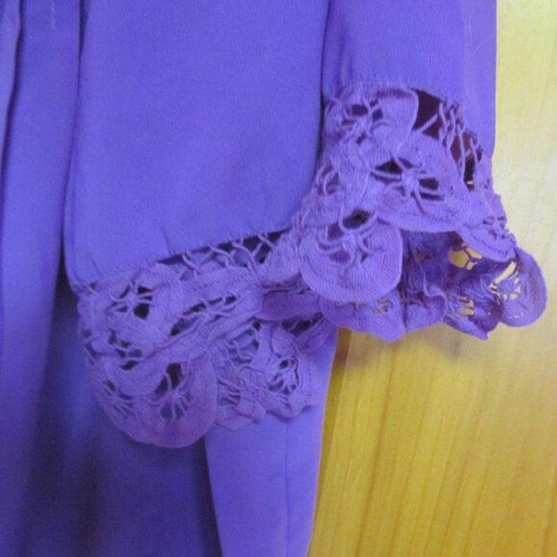 May include: A close-up of a purple garment sleeve with detailed lace trim. The lace is a darker shade of purple and forms a scalloped edge around the cuff. The fabric appears to be a soft, flowing material, typical of a blouse or dress.