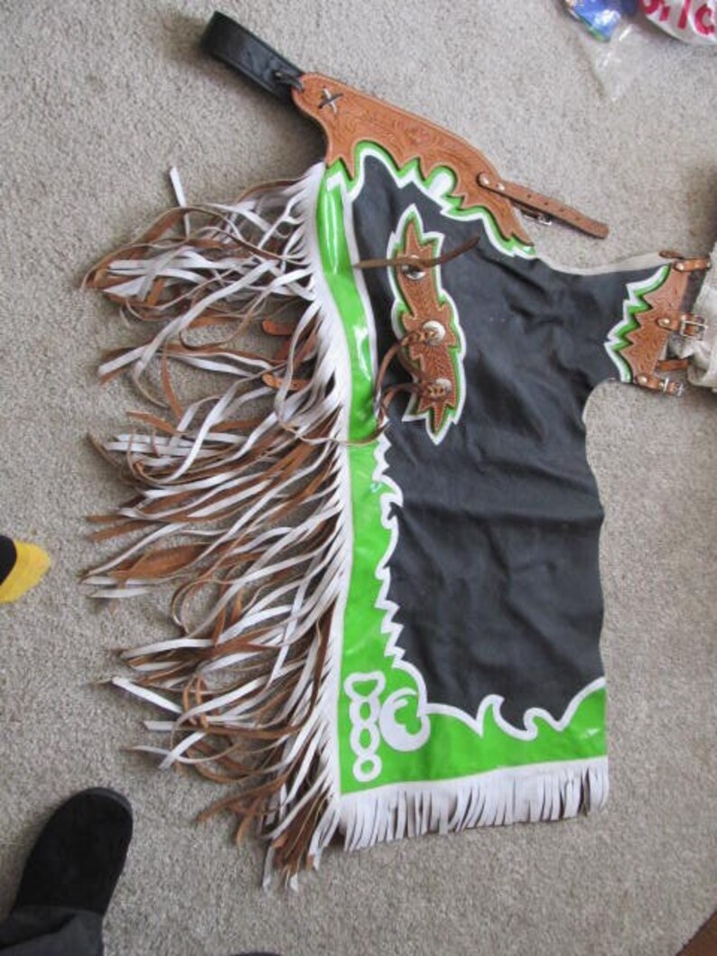 Puede incluir: Chaps de cuero negro y verde con flecos blancos y un dise&ntilde;o verde y blanco. Las chaps tienen una correa y una hebilla de cuero marr&oacute;n.