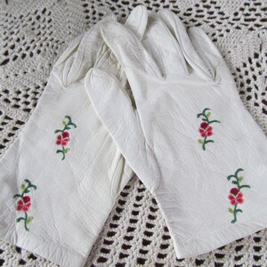 Puede incluir: Un par de guantes de cuero blanco con delicado bordado floral en tonos rojos y verdes.