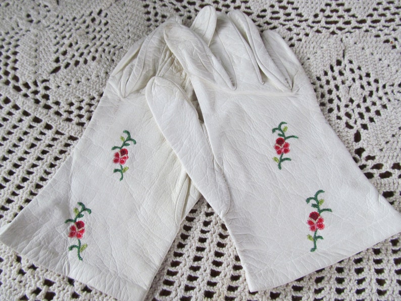 Puede incluir: Un par de guantes de cuero blanco con delicado bordado floral en tonos rojos y verdes.