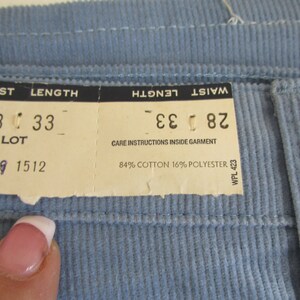 May include: A white tag with black text indicating a waist size of 28 inches and a length of 33 inches. The tag also includes the text "LOT -649 1512" and "84% COTTON 16% POLYESTER". The tag is attached to a light blue corduroy fabric.
