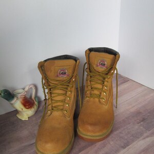 Op de afbeelding: Een paar bruine leren werkschoenen met zwarte veters. De schoenen hebben een geel logo aan de zijkant.