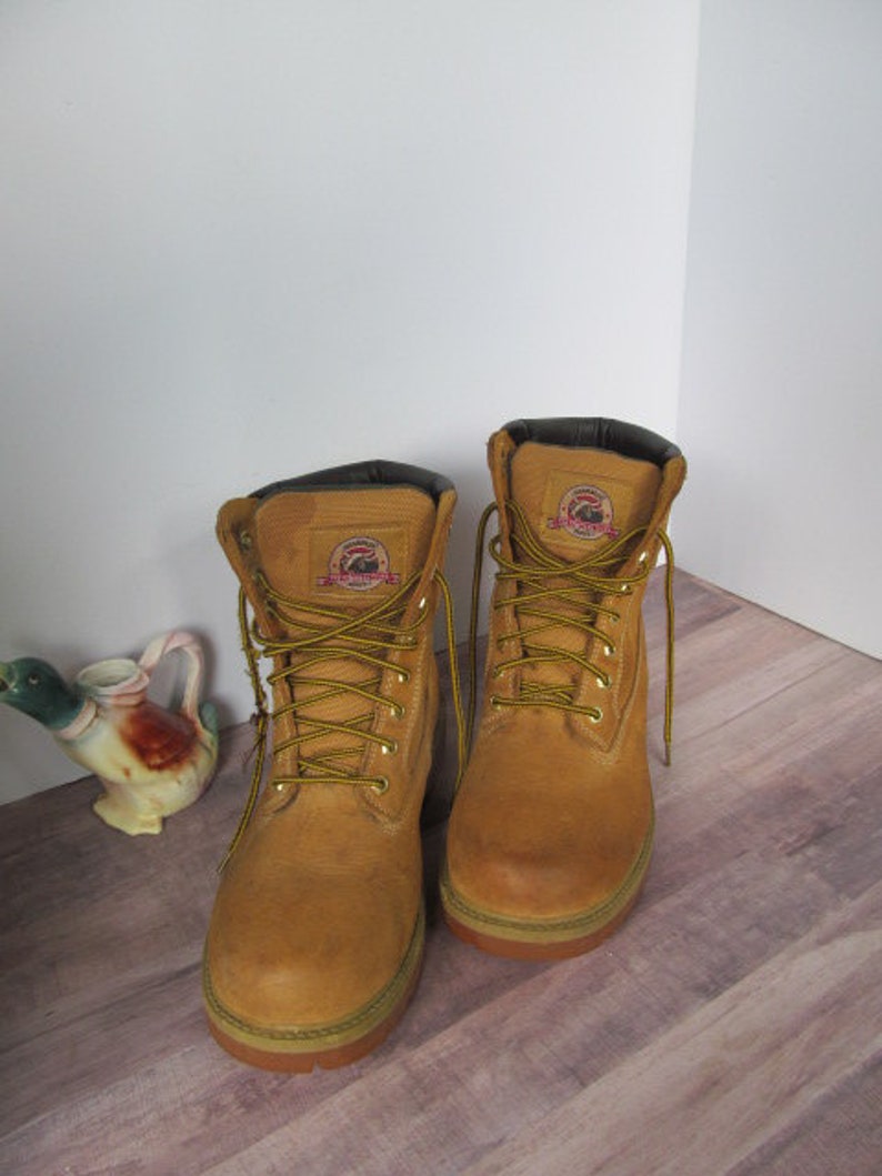 Op de afbeelding: Een paar bruine leren werkschoenen met zwarte veters. De schoenen hebben een geel logo aan de zijkant.