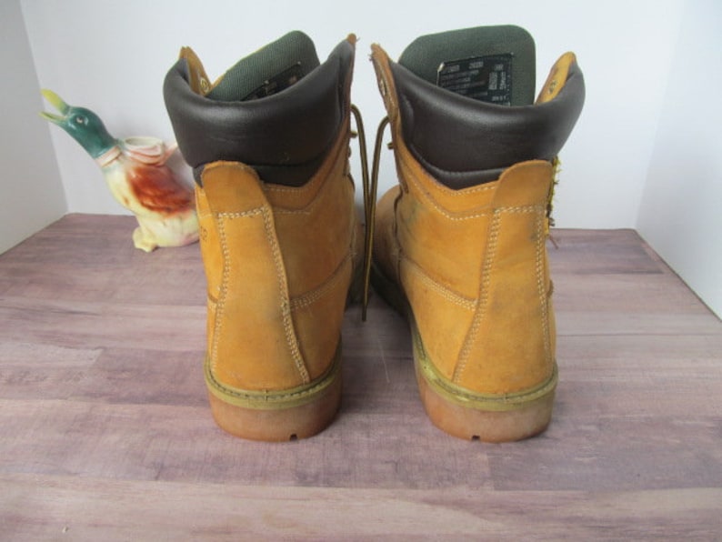 Op de afbeelding: Een paar bruine leren werkschoenen met bruine leren afwerking rond de bovenkant. De schoenen hebben veters en een dikke zool.