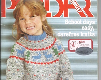 Könnte beinhalten: Ein Mädchen trägt einen grauen, blauen und roten Strickpullover mit einem Rentiermuster. Der Pullover hat einen hohen Kragen und lange Ärmel. Das Mädchen hält einen roten Schal in der Hand. Das Magazincover lautet "School days easy, carefree knits" und "Studio Knitting Machines Cone Yarns - Lessons Club Meetings".