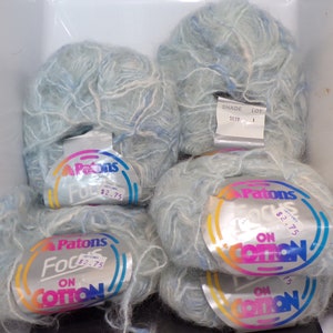 May include: Six skeins of light blue yarn with white flecks. The yarn is labeled "Patons Focus on Cotton" and has a price tag of $2.75. The yarn is in a plastic container.