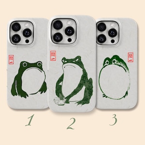 May include: Three white smartphone cases featuring green frog illustrations. The first shows a seated frog, the second a frog holding a bowl, and the third a round frog. Each case includes a small red square stamp with the Japanese text 問一.