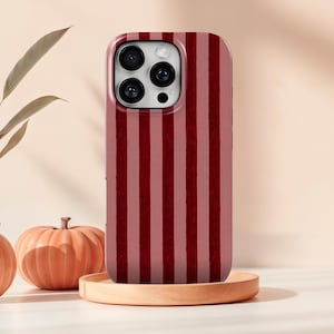 May include: A mobile phone case featuring a vertical striped design in shades of red and pink. The case rests on a wooden tray, with a pumpkin and a vase with leaves in the background.