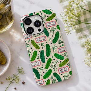 Peut inclure: Coque de téléphone avec un motif répété de cornichons verts, de brins d'aneth et le texte "you're a big dill" sur fond crème. Un bocal et une assiette de cornichons sont visibles.