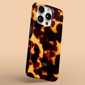 Op de afbeelding: Een telefoonhoesje met schildpadpatroon en een glanzende afwerking. De hoes heeft een donkerbruin en amberkleurig kleurenschema, met de initialen "DN" in goud. De telefoonhoes heeft een cameramodule met drie lenzen.