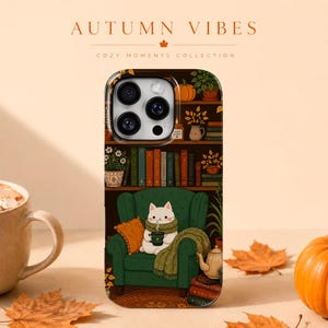 May include: A smartphone case featuring a graphic illustration of a white cat sitting in a green armchair, holding a mug, surrounded by books, plants, and a pumpkin. The text AUTUMN VIBES and COZY MOMENTS COLLECTION is printed above the phone case.