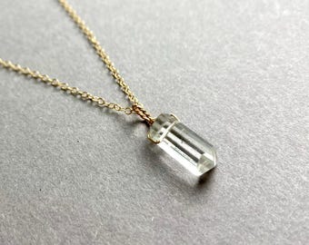 Puede incluir: Un collar de cadena dorada con un colgante de cristal de cuarzo transparente. El cristal está facetado y tiene una punta puntiaguda.