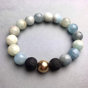 Könnte beinhalten: Ein Armband aus hellblauen, grauen und weißen Perlen mit zwei schwarzen Lavasteinen und einer goldenen Perle in der Mitte.