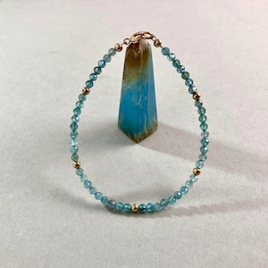 Peut inclure: Bracelet délicat composé de petites perles bleu clair facettées et de perles dorées. Le bracelet est fermé par un fermoir doré. Un obélisque en cristal bleu et marron est en arrière-plan.