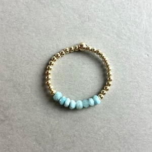 Puede incluir: Un brazalete de cuentas doradas con una fila de cuentas de piedras preciosas de color azul claro.