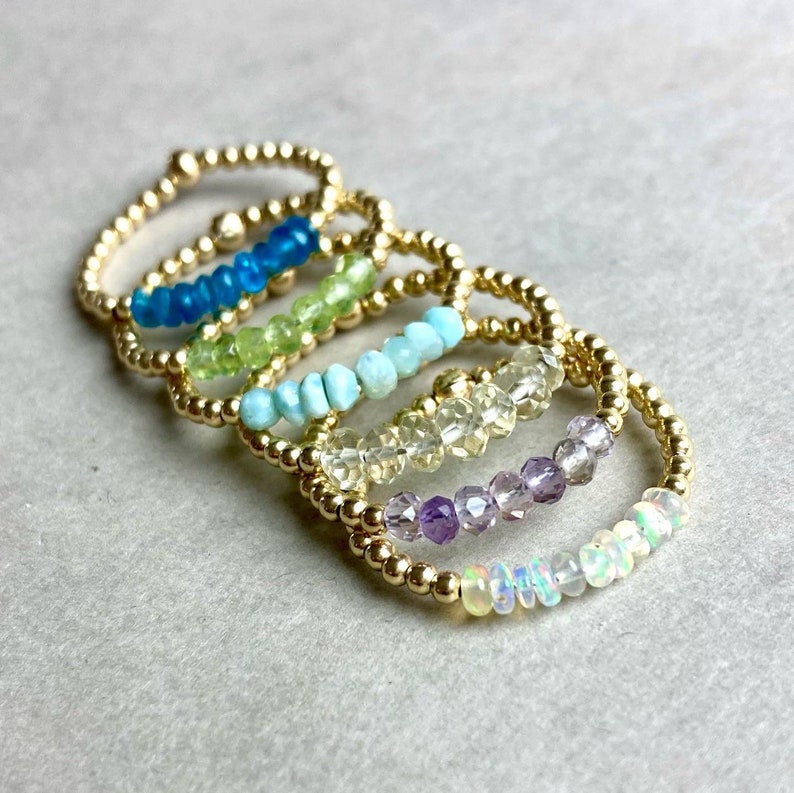 Puede incluir: Un conjunto de seis anillos de oro con piedras preciosas de diferentes colores. Los anillos est&aacute;n apilados uno encima del otro, con las piedras preciosas mirando hacia afuera. Las piedras preciosas son azules, verdes, turquesas, amarillas, moradas y blancas.