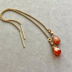 May include: Gold-coloured chain earrings with a drop design. Each earring features a delicate chain that leads to a small, teardrop-shaped, orange-red gemstone. The earrings have a simple, elegant style.