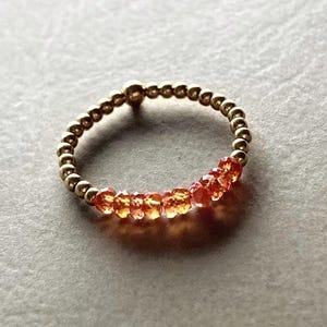 May include: A gold-coloured beaded ring with a row of orange gemstones. The ring features small, round beads forming the band, with a cluster of faceted orange stones set in the centre. The gemstones add a pop of colour to the jewellery.