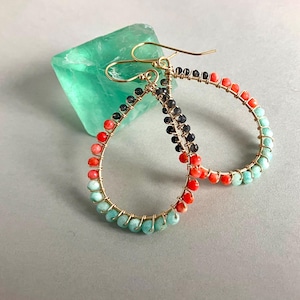 May include: A pair of teardrop-shaped hoop earrings crafted from gold-toned wire wrapped with small faceted beads in shades of coral, turquoise, and dark grey. The earrings feature a hook-style closure and are arranged in a circular pattern.