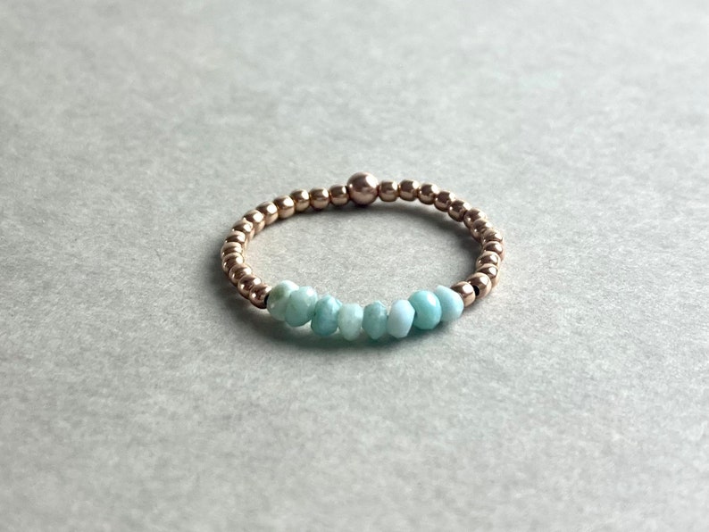 Puede incluir: Un delicado anillo de oro con una fila de cuentas de piedras preciosas de color azul claro.