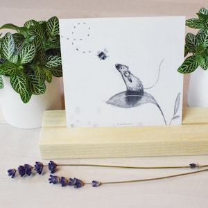 May include: A black and white illustration of a mouse on a leaf with a bee flying nearby. The illustration is printed on a white card and displayed on a wooden stand.