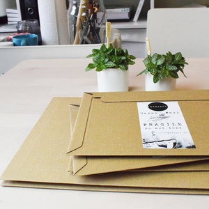 May include: Three brown cardboard boxes stacked on top of each other. The top box has a white label that says "Happy Mail" and "Fragile Do not bend". Two small potted plants with green leaves are in the background.
