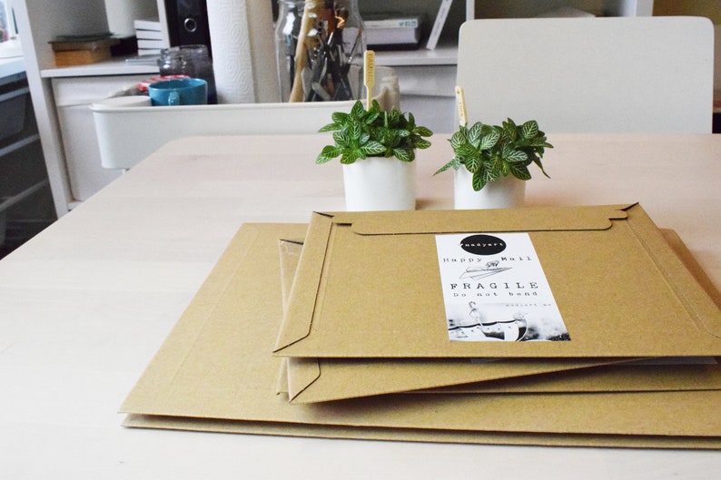 May include: Three brown cardboard boxes stacked on top of each other. The top box has a white label that says "Happy Mail" and "Fragile Do not bend". Two small potted plants with green leaves are in the background.