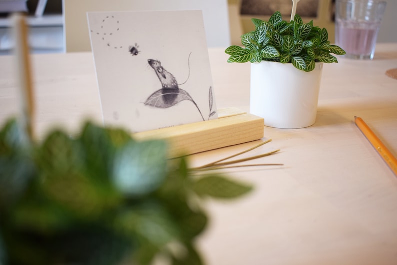 May include: A black and white drawing of a mouse on a leaf, framed in a wooden stand. The drawing is on a white card with a light pink background. There is a small potted plant with green leaves in the background.