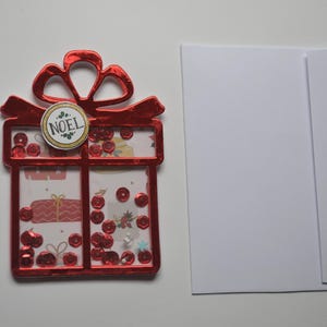 May include: A red Christmas gift-shaped shaker card with a bow and the word "NOEL" on a circular tag. The card is filled with red sequins and festive decorations. A white envelope is next to the card.