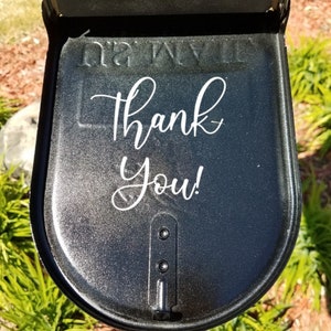 Puede incluir: Una puerta de buz&oacute;n de metal negro con el texto Thank You! en letra cursiva blanca sobre la superficie. La puerta tiene un acabado met&aacute;lico brillante e incluye un mecanismo de cierre vertical central con dos remaches visibles.