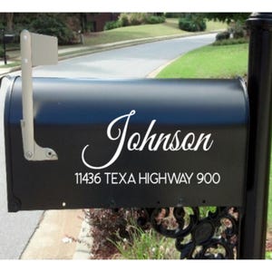 May include: A matte black metal mailbox featuring a white vinyl decal with the name Johnson in a cursive script font, followed by the address 11436 TEXA HIGHWAY 900 in a clean sans-serif typeface, mounted on a black post with a side flag.