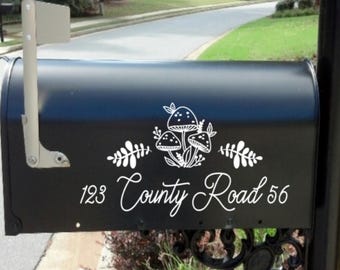 May include: A matte black metal mailbox featuring a white vinyl decal of three mushrooms surrounded by fern-like leaves. Below the graphic, the text 123 County Road 56 is displayed in a white cursive font. The mailbox has a silver flag on the side.