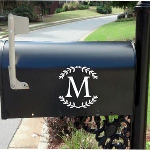 May include: A black mailbox with a white wreath design and the letter "M". The mailbox has a silver flag and is mounted on a black post. The background shows a residential street and green grass.