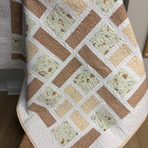 May include: A baby quilt with a patchwork design. The quilt features squares of light green fabric with woodland animal prints, alternating with tan and cream-coloured blocks. The quilt is bordered with white fabric and has a quilted pattern.