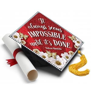 Puede incluir: Un birrete de graduación con la parte superior roja brillante, con la cita "It always seems impossible until it's done" de Nelson Mandela. El birrete está decorado con flores blancas, mariposas doradas y una borla dorada. También hay un diploma enrollado con una cinta roja.