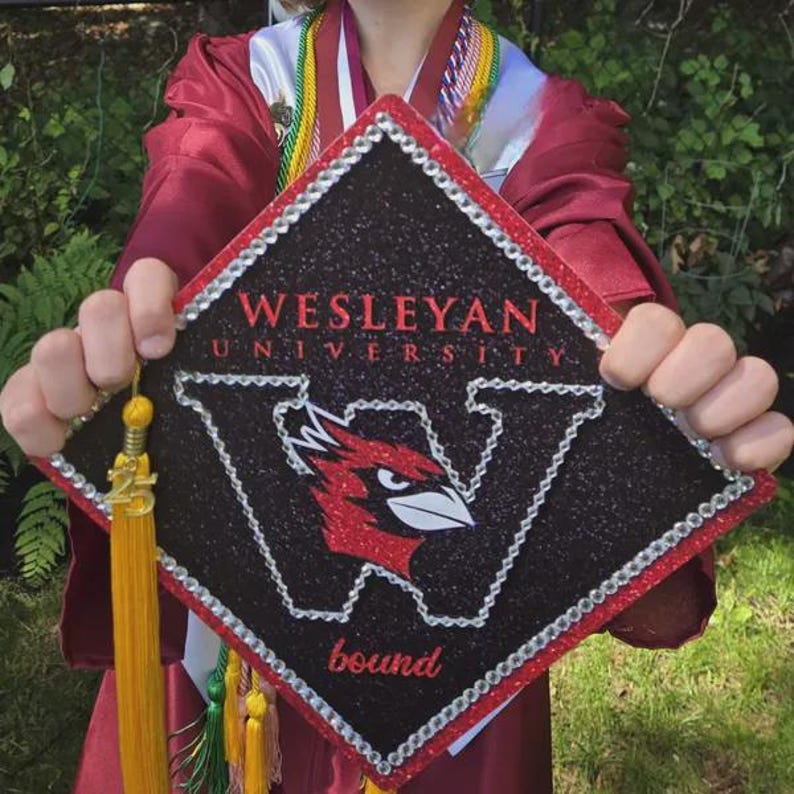 A person wearing a red graduation cap and gown holds up a black and red graduation cap with the text "Wesleyan University" and a bird logo.
