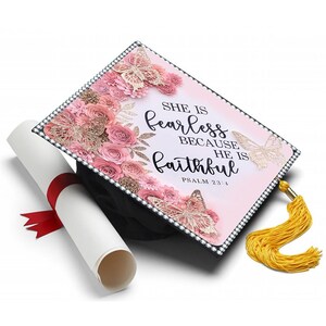 May include: A black graduation cap decorated with pink roses, butterflies, and the text "She is fearless because He is faithful, Psalm 23:4." A rolled diploma with a red ribbon and a gold tassel are also present.