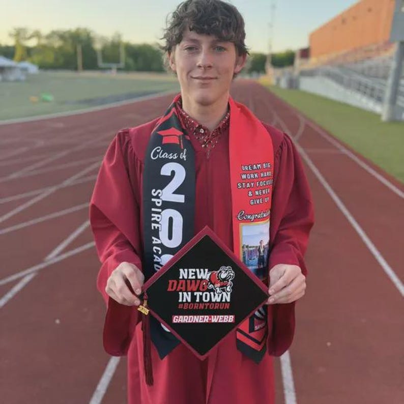 A smiling young man in a red graduation gown holding a black graduation cap with text and graphics.