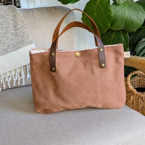Peut inclure: Un sac fourre-tout en toile marron avec des poignées en cuir marron et des rivets en laiton. Le sac a une fermeture à bouton et un intérieur à motifs. Le sac est posé sur une chaise rembourrée grise.