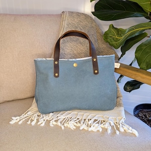 May include: A blue tote bag with brown leather handles and gold rivets. The bag is sitting on a beige chair with a fringed blanket. A houseplant is in the background.