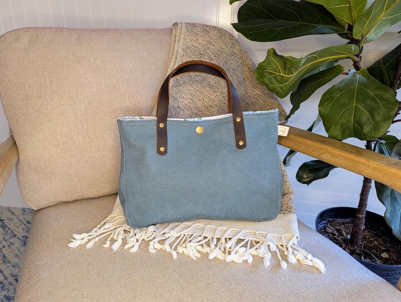 May include: A blue tote bag with brown leather handles and gold rivets. The bag is sitting on a beige chair with a fringed blanket. A houseplant is in the background.